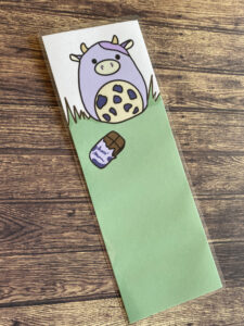 Marque page d’un dessin d’une mignonne petite vache violette accompagnée d’une tablette de chocolat dont l’emballage est aussi violet 