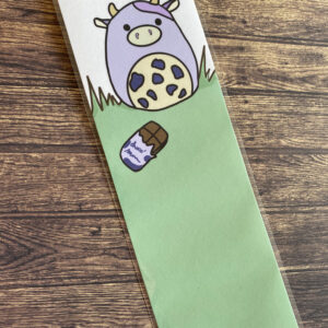 Marque page d’un dessin d’une mignonne petite vache violette accompagnée d’une tablette de chocolat dont l’emballage est aussi violet